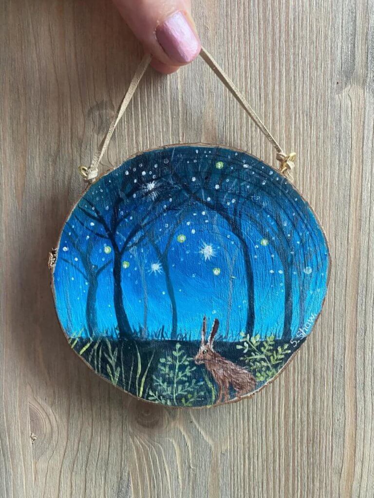 A hand painted wooden decoration depicting a rabbit in the woods surrounded by stars.